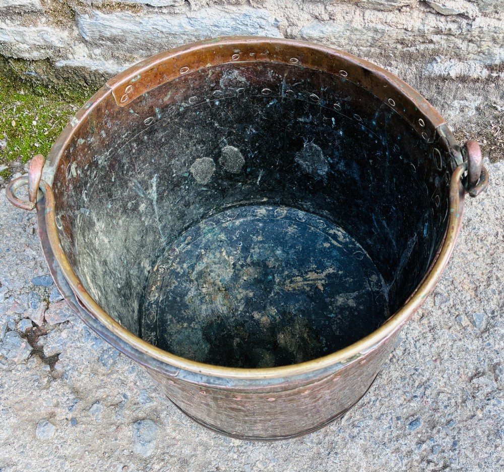 An antique cylindrical copper bucket featuring heavy copper rivets along the upper rim band and a metal swing handle. View V