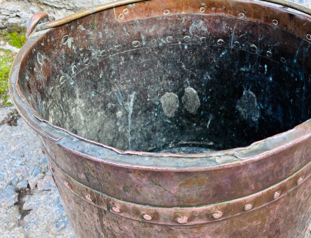 An antique cylindrical copper bucket featuring heavy copper rivets along the upper rim band and a metal swing handle. View VI