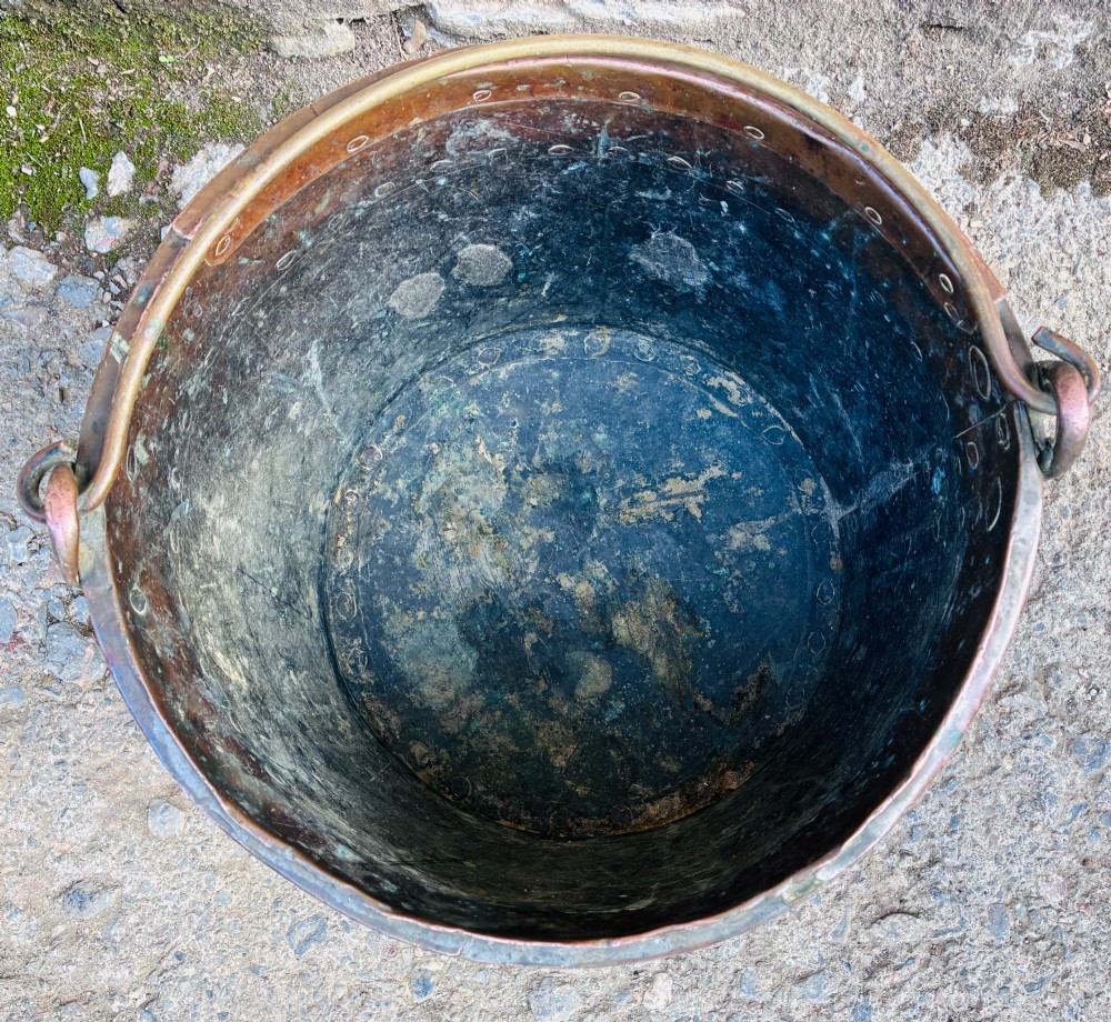 An antique cylindrical copper bucket featuring heavy copper rivets along the upper rim band and a metal swing handle. View VII