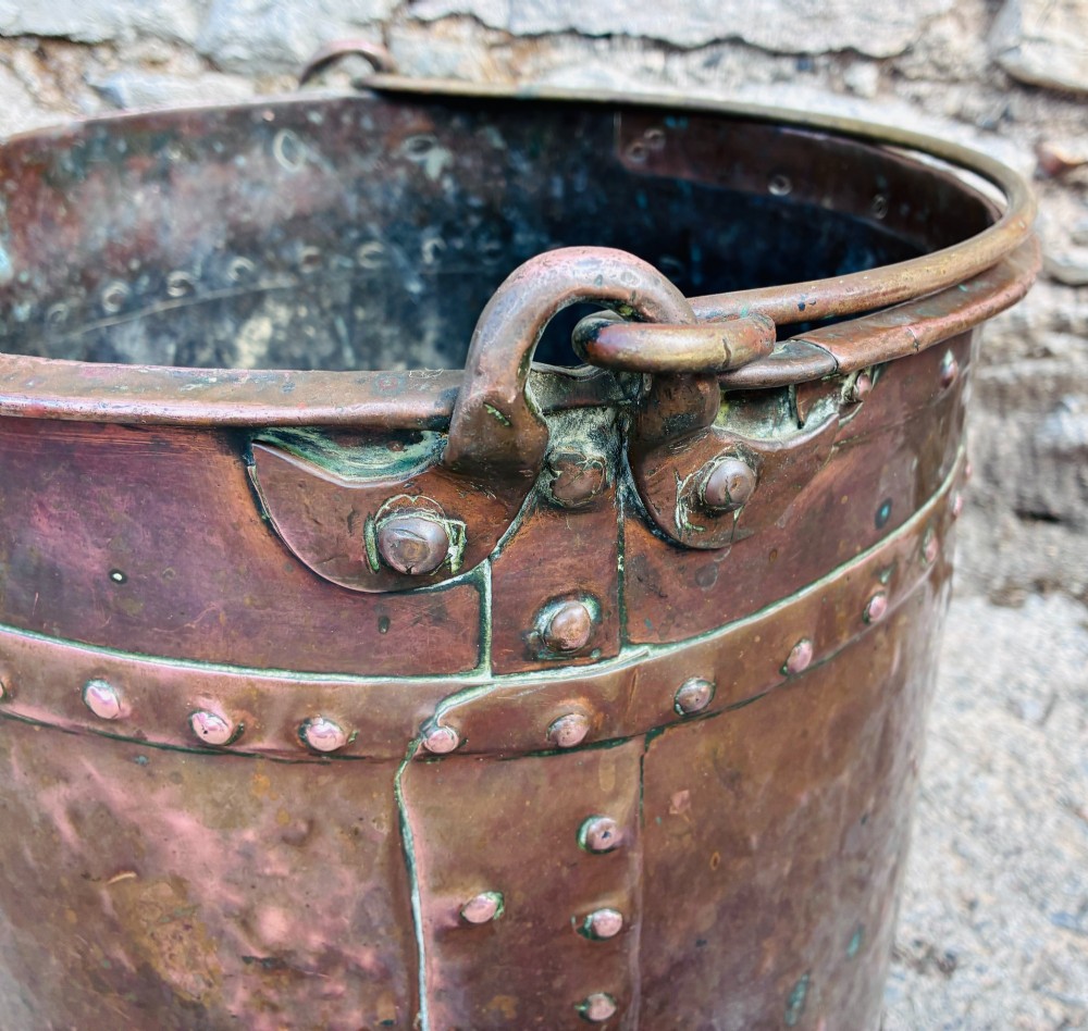 An antique cylindrical copper bucket featuring heavy copper rivets along the upper rim band and a metal swing handle. View IX