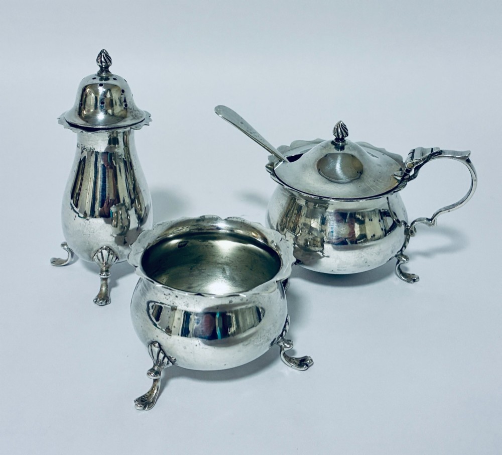 A three-piece silver cruet set comprising a pepper caster, open salt cellar, and mustard pot with a spoon, each resting on shell-adorned feet.