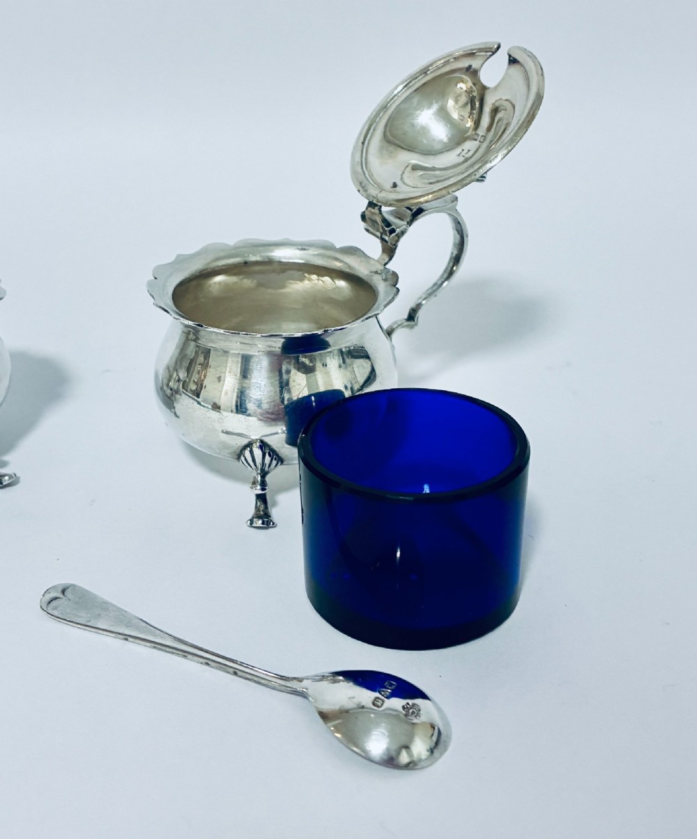 A three-piece silver cruet set comprising a pepper caster, open salt cellar, and mustard pot with a spoon, each resting on shell-adorned feet. View IV