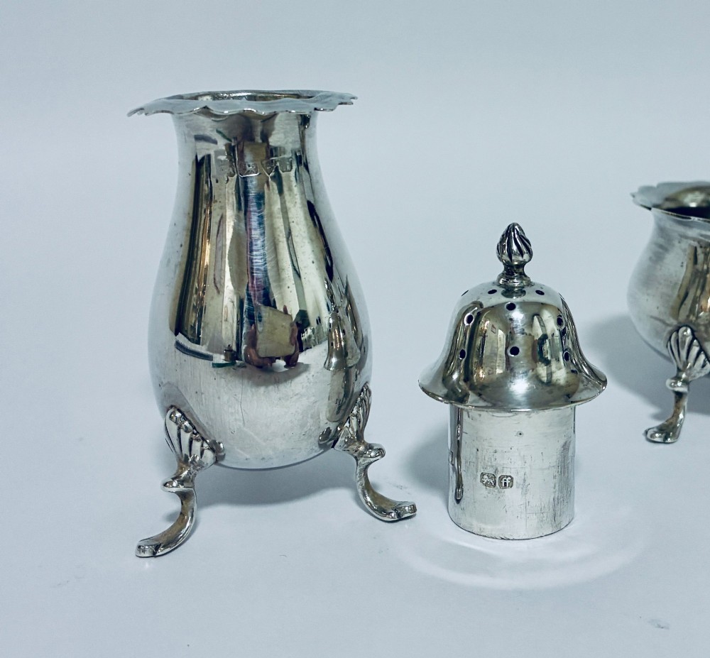 A three-piece silver cruet set comprising a pepper caster, open salt cellar, and mustard pot with a spoon, each resting on shell-adorned feet. View VI