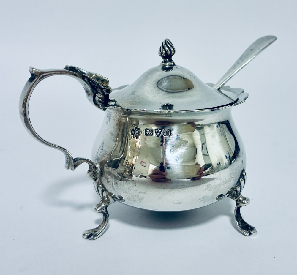 A three-piece silver cruet set comprising a pepper caster, open salt cellar, and mustard pot with a spoon, each resting on shell-adorned feet. View IX
