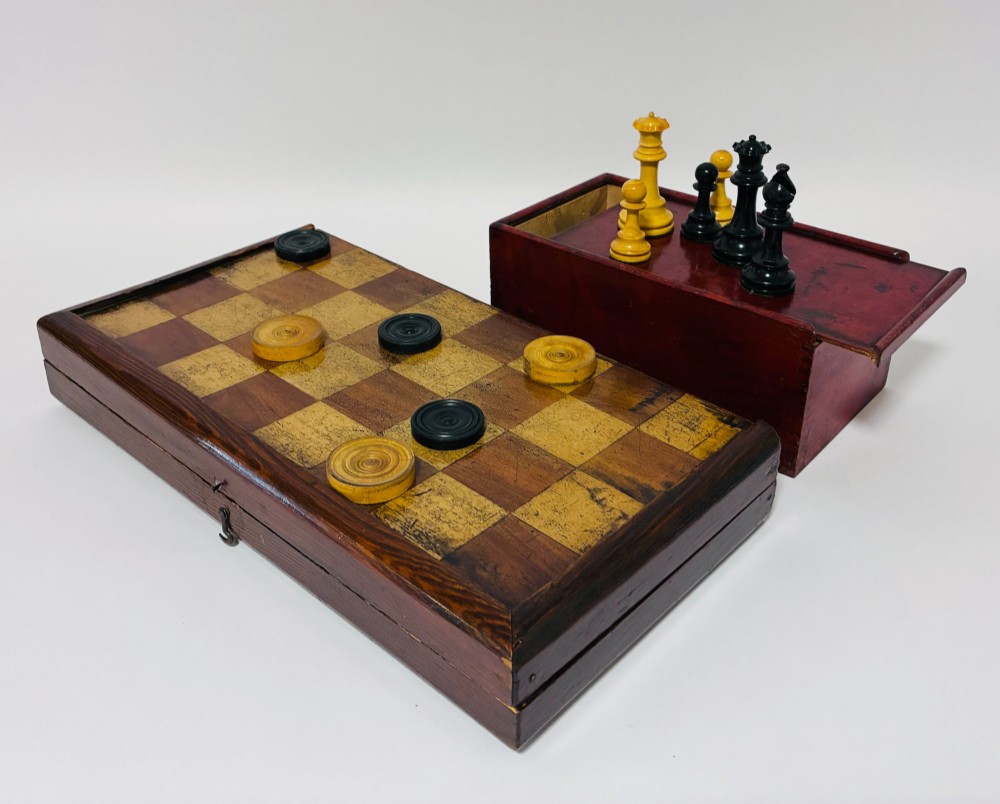 An antique hinged wooden folding games board with a checkerboard surface, accompanied by turned draughts counters and a sliding wooden box containing chess pieces.