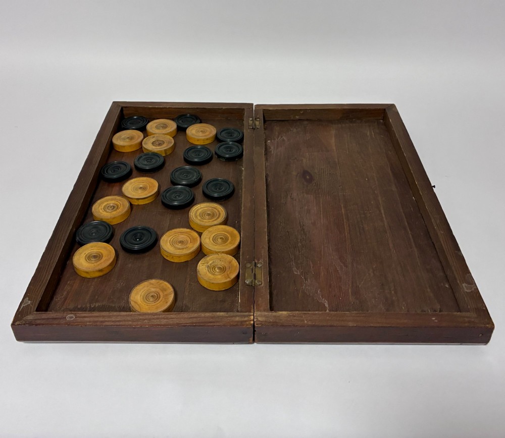 An antique hinged wooden folding games board with a checkerboard surface, accompanied by turned draughts counters and a sliding wooden box containing chess pieces. View IV