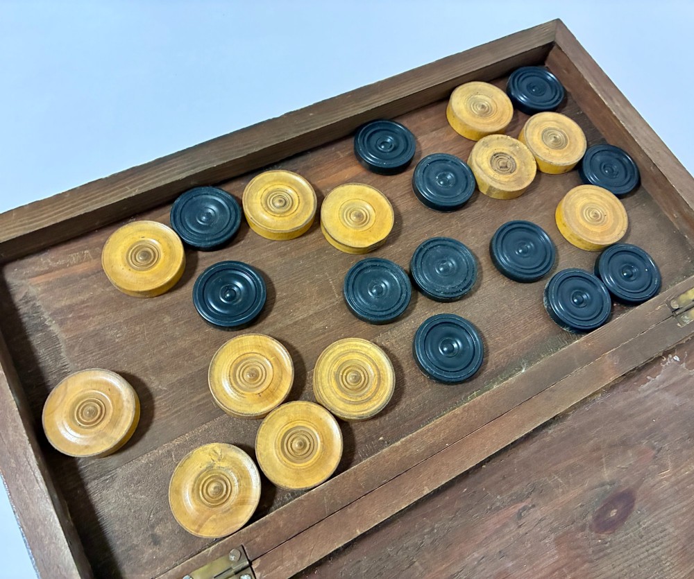 An antique hinged wooden folding games board with a checkerboard surface, accompanied by turned draughts counters and a sliding wooden box containing chess pieces. View V