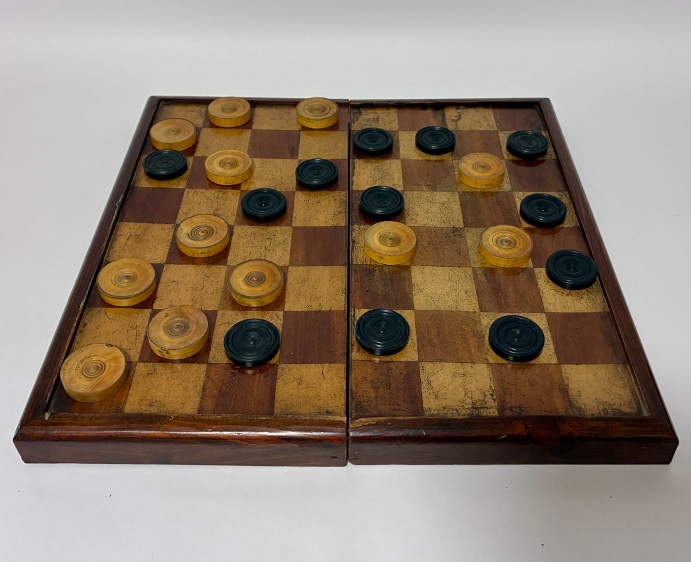 An antique hinged wooden folding games board with a checkerboard surface, accompanied by turned draughts counters and a sliding wooden box containing chess pieces. View VI