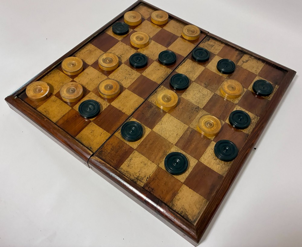 An antique hinged wooden folding games board with a checkerboard surface, accompanied by turned draughts counters and a sliding wooden box containing chess pieces. View VII
