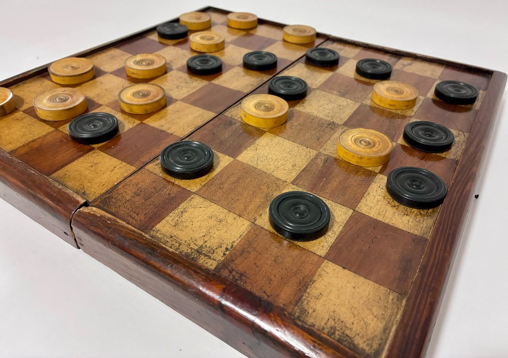 An antique hinged wooden folding games board with a checkerboard surface, accompanied by turned draughts counters and a sliding wooden box containing chess pieces. View VIII