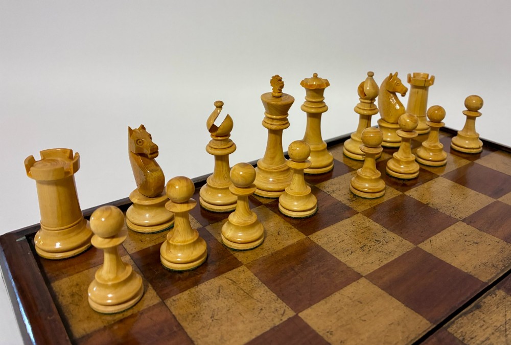 An antique hinged wooden folding games board with a checkerboard surface, accompanied by turned draughts counters and a sliding wooden box containing chess pieces. View XII