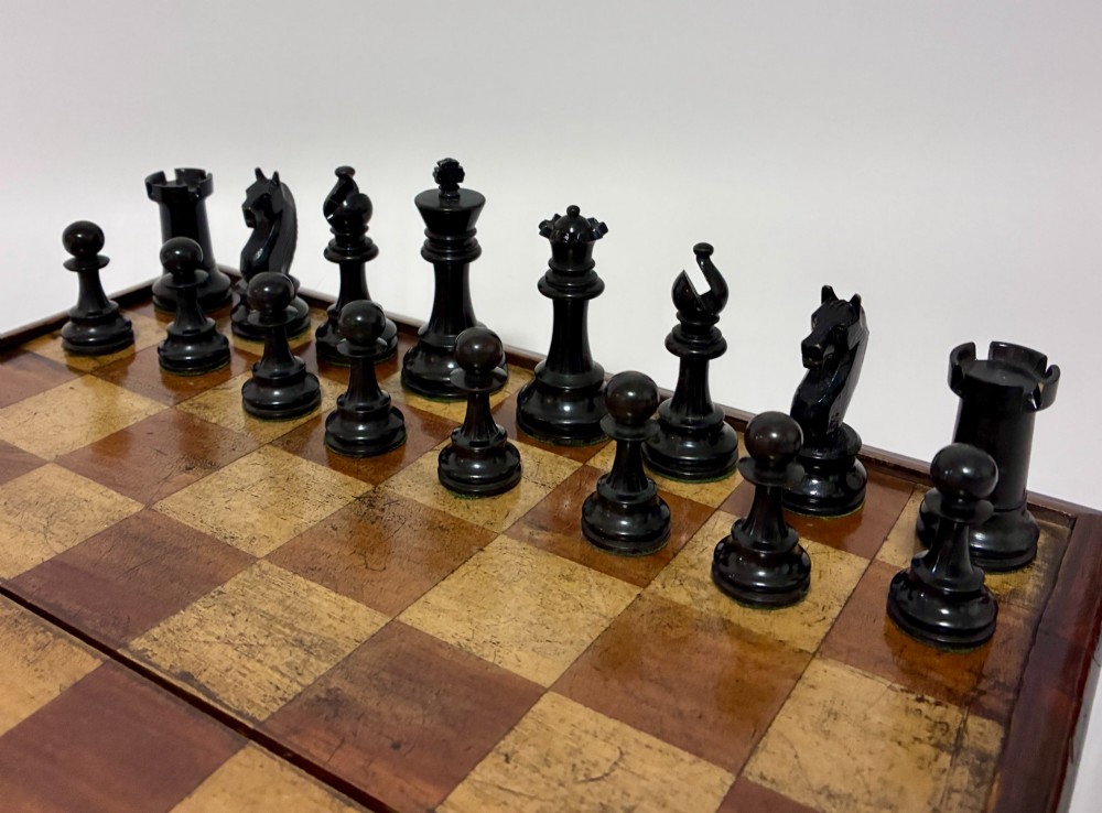 An antique hinged wooden folding games board with a checkerboard surface, accompanied by turned draughts counters and a sliding wooden box containing chess pieces. View XIV