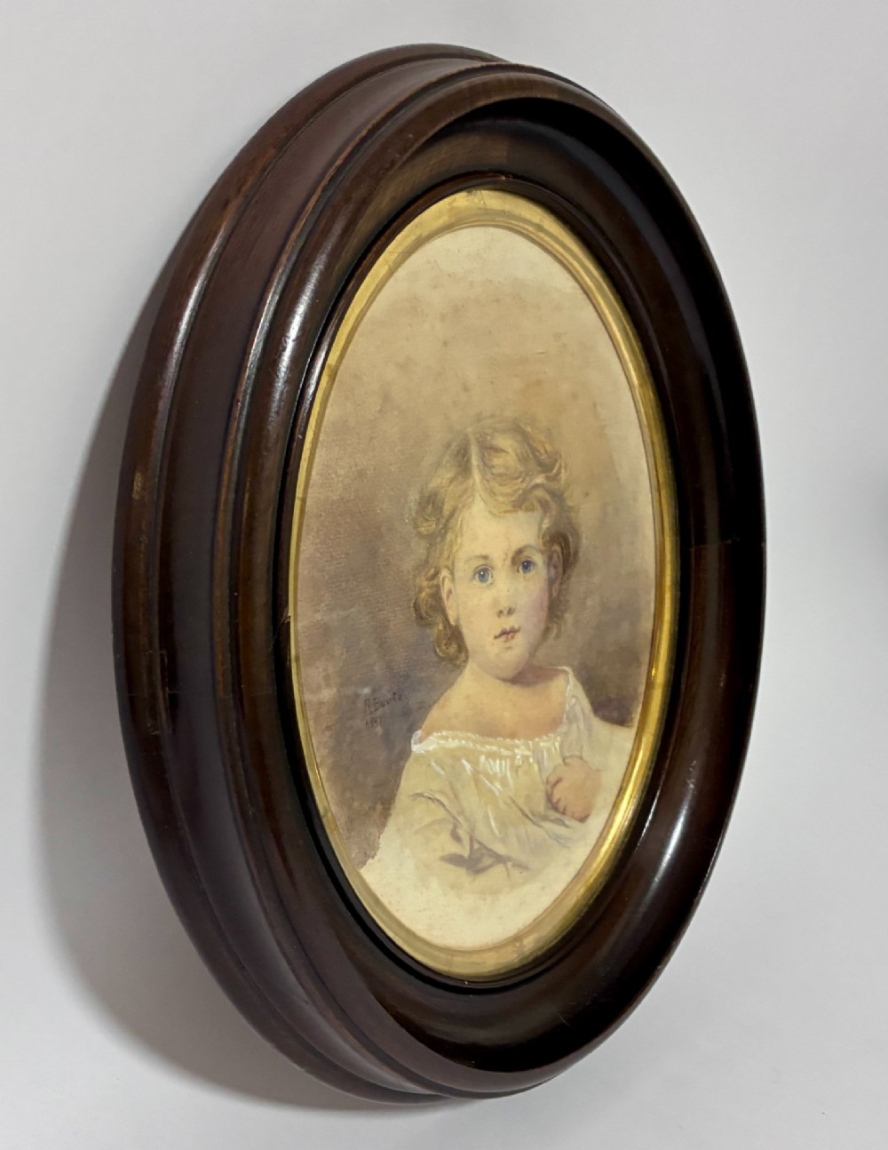 An oval Edwardian watercolour portrait depicting a young child with blonde curls, set inside a dark wooden frame with a gilt inner trim. View IV