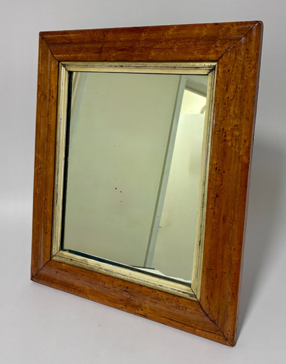 An antique rectangular birds eye maple frame featuring an inner gilt slip rim surrounding a mirror glass plate, supported by an easel stand.