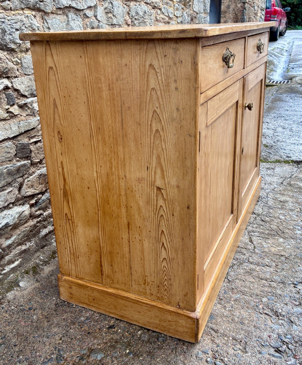 A nineteenth-century stripped pine sideboard featuring two upper drawers with ornate brass handles above two panelled cupboard doors on a solid plinth base. View IV