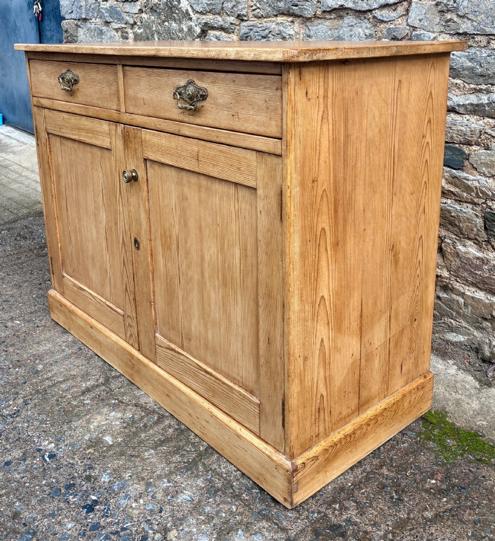 A nineteenth-century stripped pine sideboard featuring two upper drawers with ornate brass handles above two panelled cupboard doors on a solid plinth base. View V
