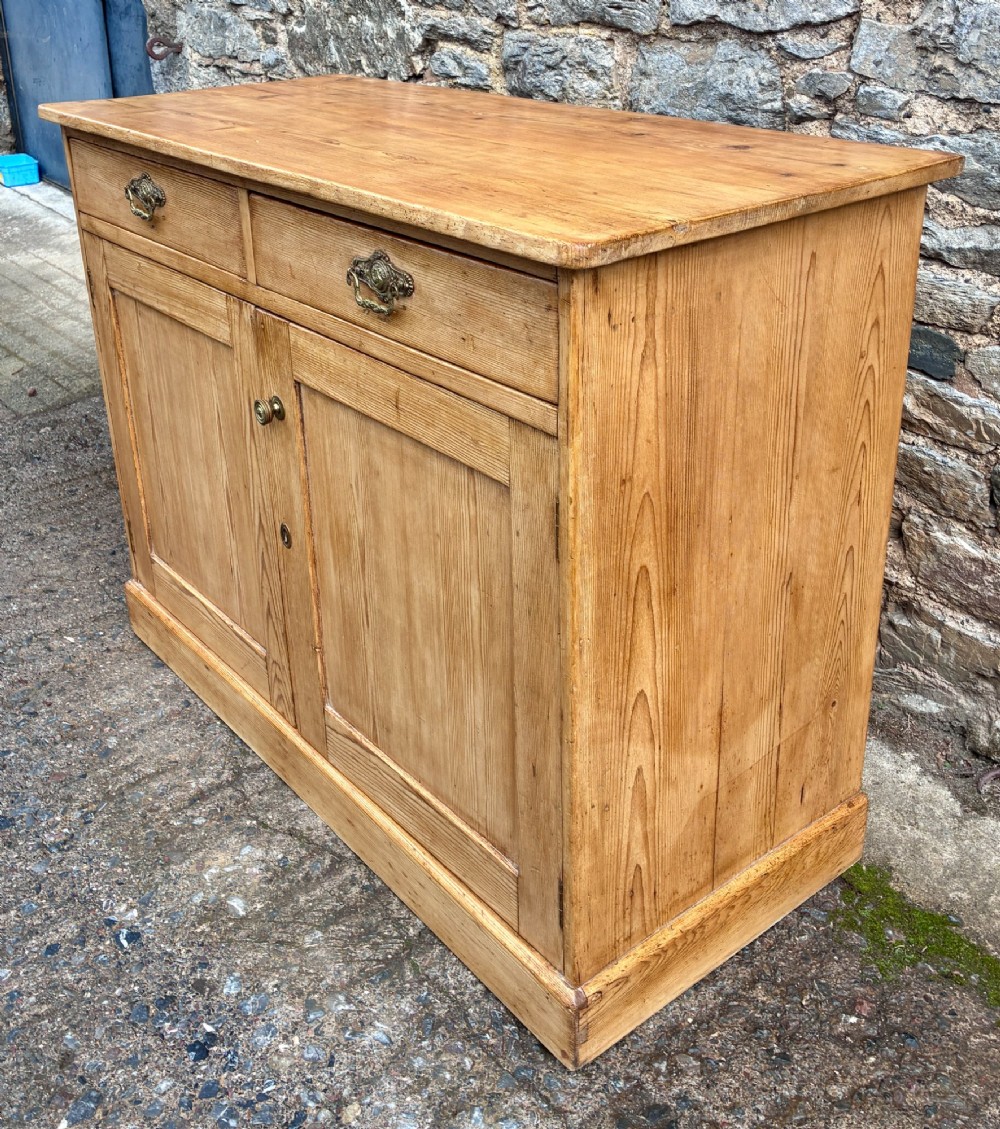 A nineteenth-century stripped pine sideboard featuring two upper drawers with ornate brass handles above two panelled cupboard doors on a solid plinth base. View VI