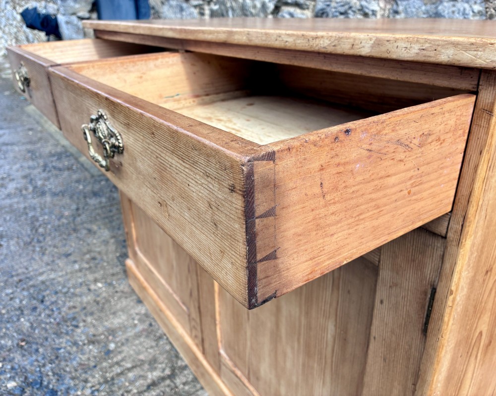 A nineteenth-century stripped pine sideboard featuring two upper drawers with ornate brass handles above two panelled cupboard doors on a solid plinth base. View X