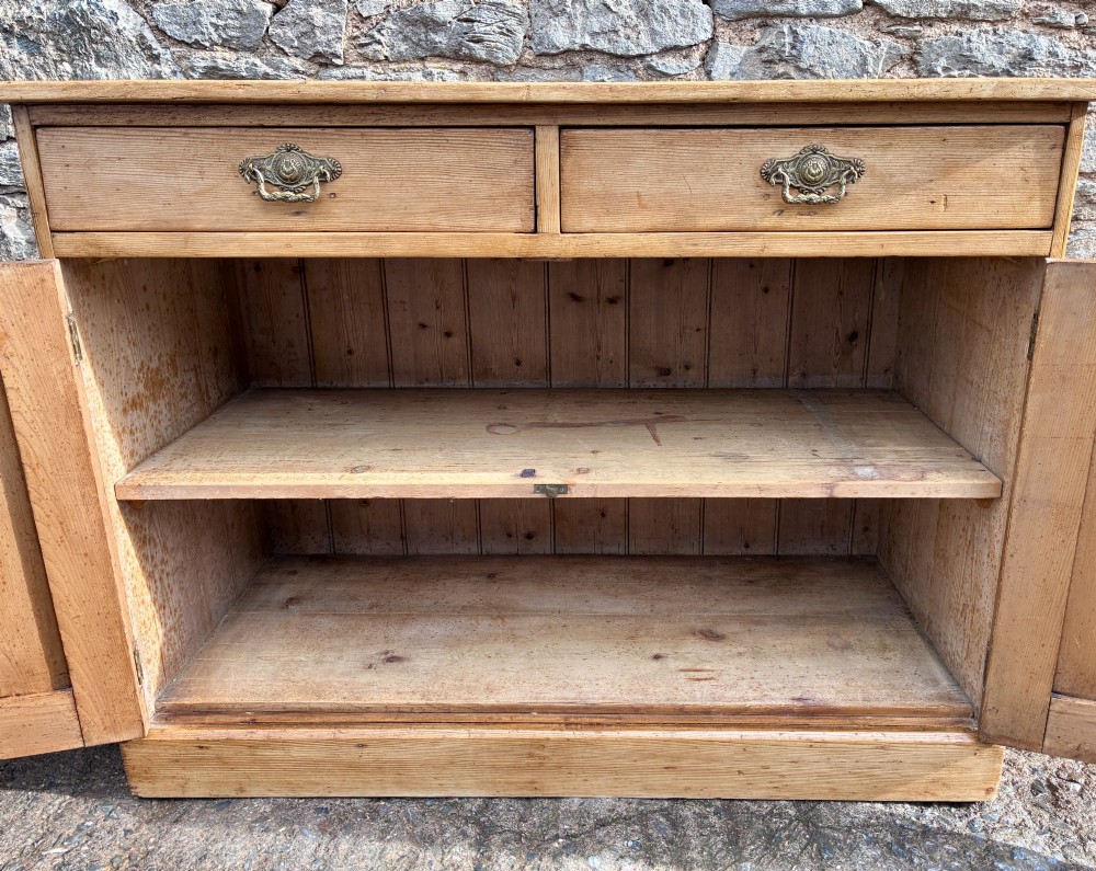 A nineteenth-century stripped pine sideboard featuring two upper drawers with ornate brass handles above two panelled cupboard doors on a solid plinth base. View XI