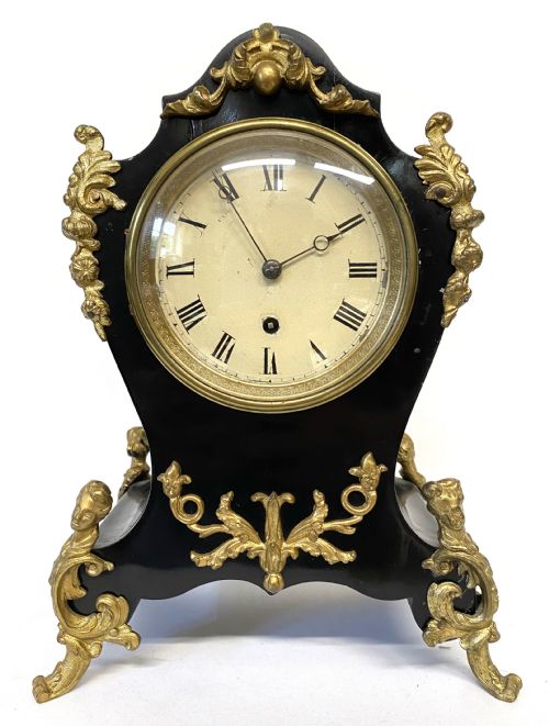 fig 1. An antique ebonised wooden mantel clock featuring ornate cast ormolu mounts, a white dial with Roman numerals, and scrolled gilt brass feet.