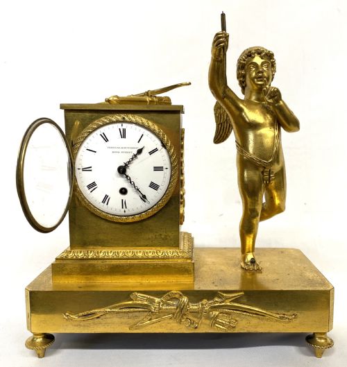 fig 1. An antique gilt bronze mantel clock featuring a white enamel dial alongside a standing Cupid figure holding an arrow, set upon a decorative gilded plinth base.