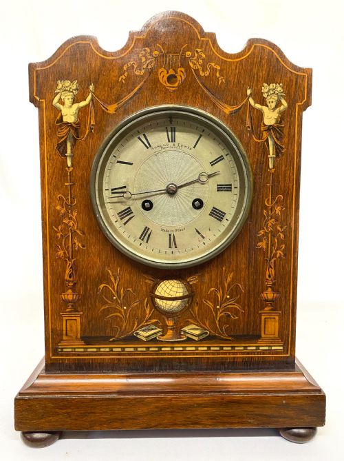 fig 1. An antique rosewood mantel clock featuring intricate marquetry inlays of figures, swags, and a globe surrounding a circular dial with Roman numerals.
