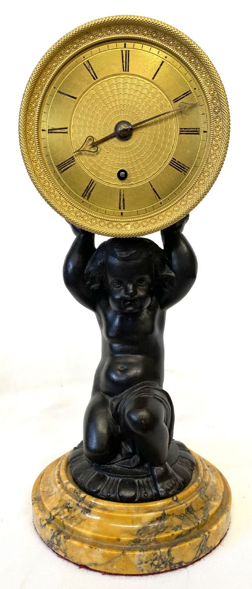 fig 2. An antique gilt brass and dark patinated bronze mantel clock depicting a kneeling cherub supporting a circular drum dial above a veined yellow marble base.