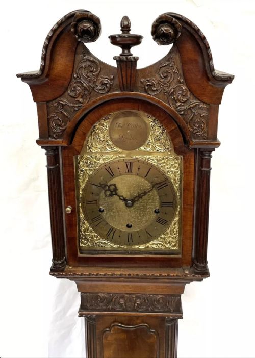 fig 3. An antique mahogany grandmother clock featuring a swan-neck pediment, carved scrollwork details, fluted columns, and an ornate brass dial with Roman numerals.