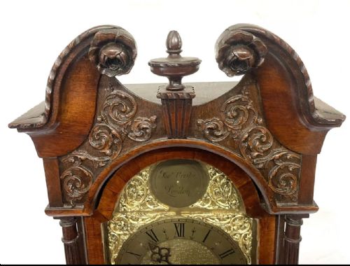 fig 5. An antique mahogany grandmother clock featuring a swan-neck pediment, carved scrollwork details, fluted columns, and an ornate brass dial with Roman numerals.