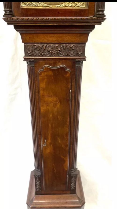 fig 6. An antique mahogany grandmother clock featuring a swan-neck pediment, carved scrollwork details, fluted columns, and an ornate brass dial with Roman numerals.