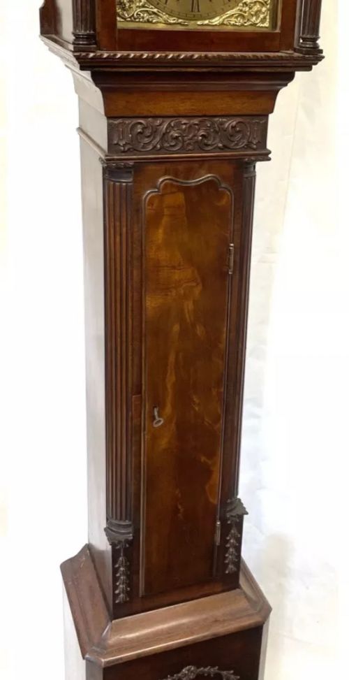 fig 7. An antique mahogany grandmother clock featuring a swan-neck pediment, carved scrollwork details, fluted columns, and an ornate brass dial with Roman numerals.