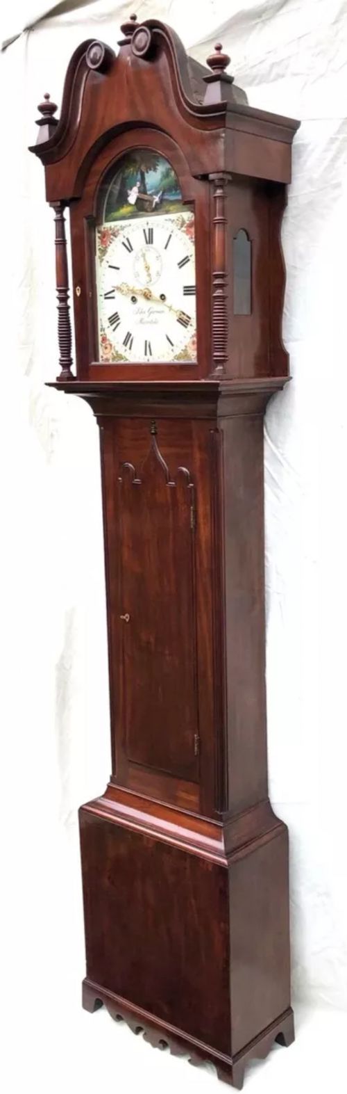 fig 1. An antique mahogany longcase clock featuring a swan-neck pediment, brass finials, a painted arch dial with a rocking seesaw automaton, and a gothic-arched trunk door.