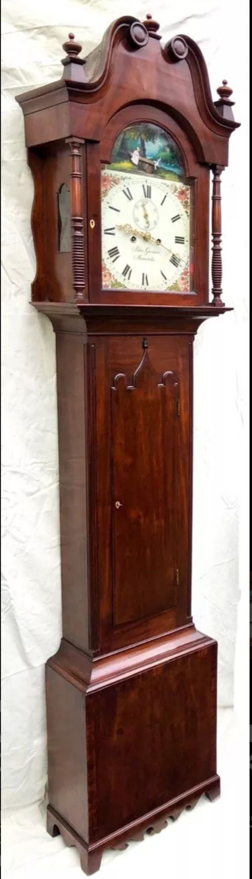 fig 2. An antique mahogany longcase clock featuring a swan-neck pediment, brass finials, a painted arch dial with a rocking seesaw automaton, and a gothic-arched trunk door.