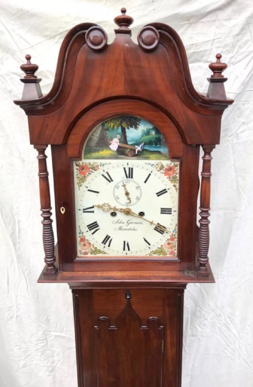 fig 3. An antique mahogany longcase clock featuring a swan-neck pediment, brass finials, a painted arch dial with a rocking seesaw automaton, and a gothic-arched trunk door.