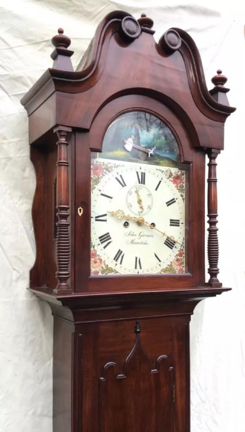 fig 4. An antique mahogany longcase clock featuring a swan-neck pediment, brass finials, a painted arch dial with a rocking seesaw automaton, and a gothic-arched trunk door.