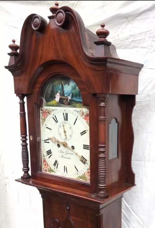 fig 5. An antique mahogany longcase clock featuring a swan-neck pediment, brass finials, a painted arch dial with a rocking seesaw automaton, and a gothic-arched trunk door.