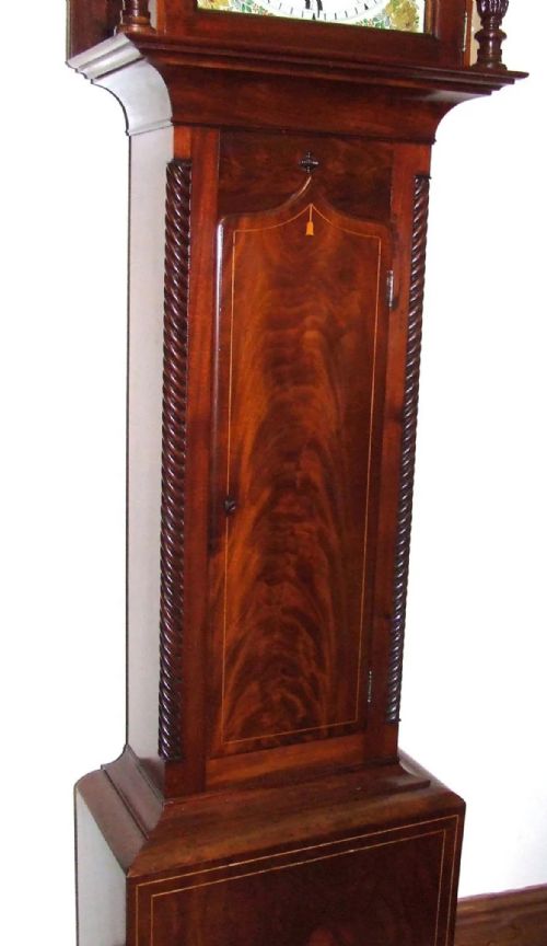 fig 6. An antique inlaid mahogany longcase clock featuring a swan-neck pediment with finials, a painted arch dial, and twisted side columns.