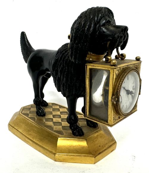 fig 2. A French gilt and patinated bronze timepiece featuring a standing dog figure holding a square glass clock case in its mouth on a gilded base.