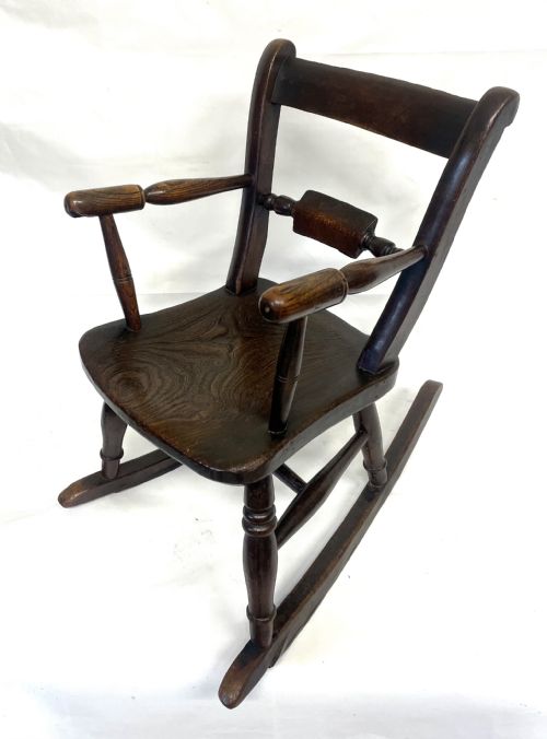 fig 1. A dark brown wooden childs or dolls rocking armchair featuring turned legs, curved armrests, and smooth wooden rockers.