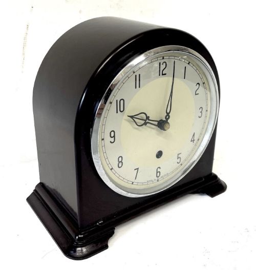 fig 2. A dark brown domed Bakelite mantel clock with a round cream dial, black Arabic numerals, spade hands, and a broad arrow symbol.