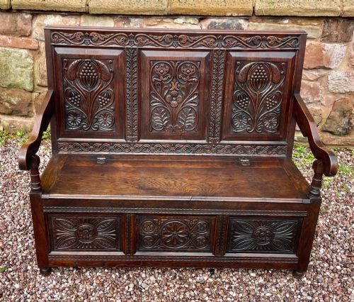 fig 2. A dark carved oak box settle featuring relief floral panels on the backrest, curved armrests, and a lift-up storage seat.