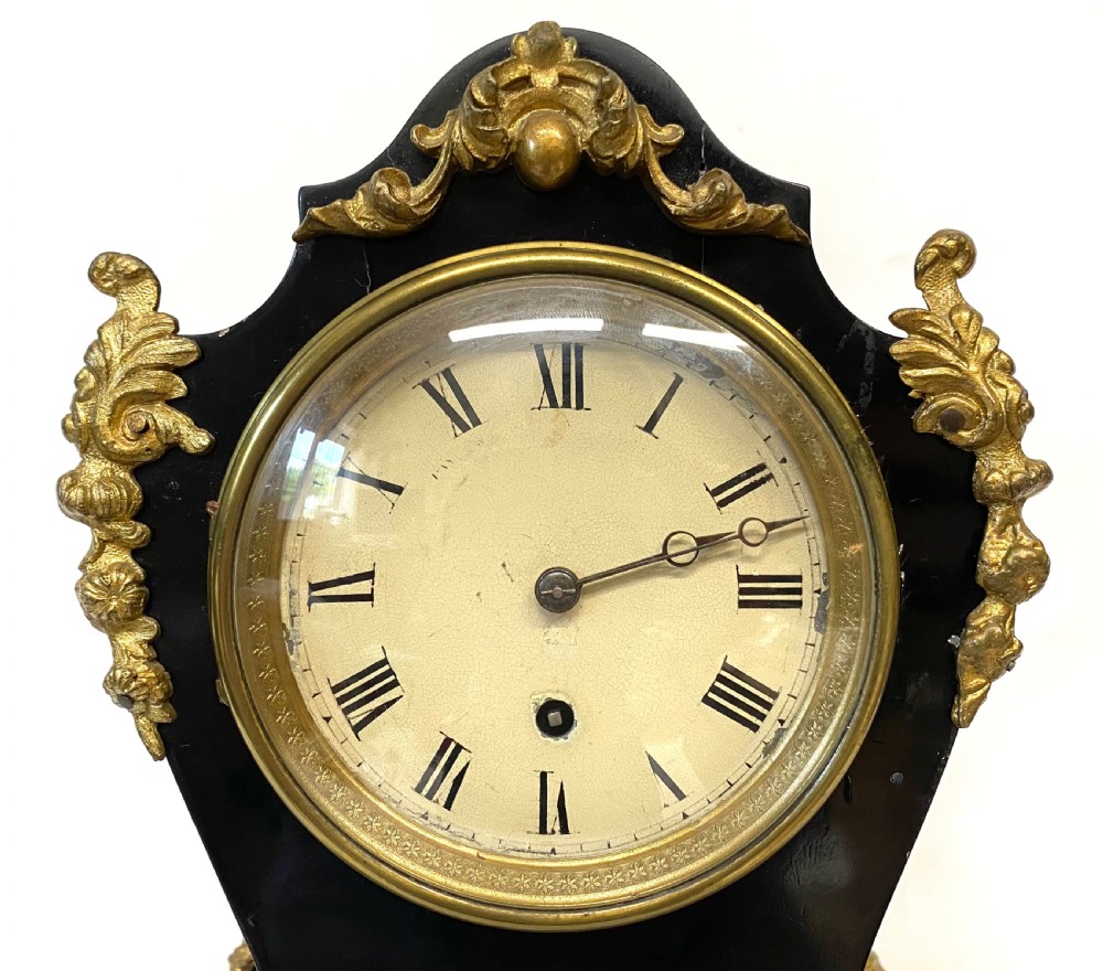An antique ebonised wooden mantel clock featuring ornate cast ormolu mounts, a white dial with Roman numerals, and scrolled gilt brass feet. View V
