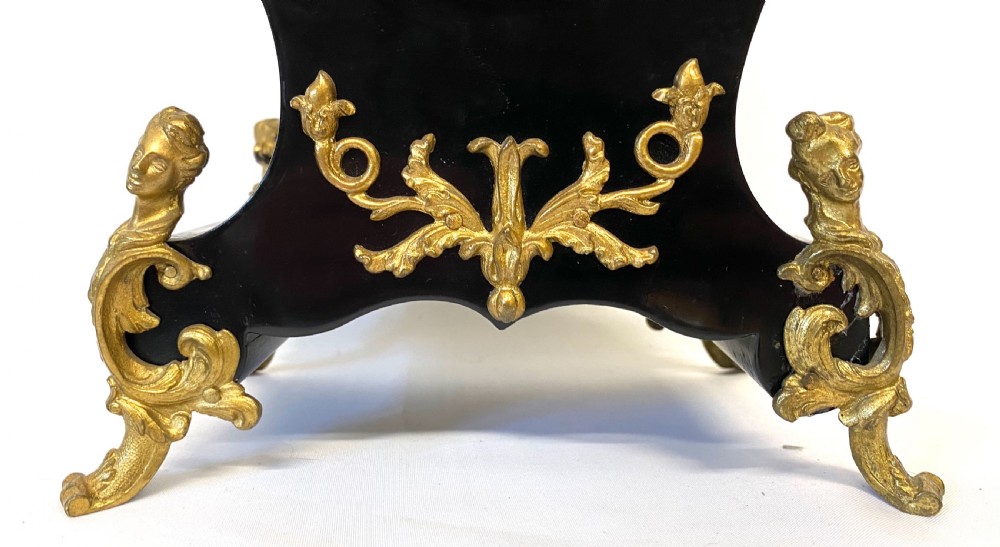 An antique ebonised wooden mantel clock featuring ornate cast ormolu mounts, a white dial with Roman numerals, and scrolled gilt brass feet. View VII