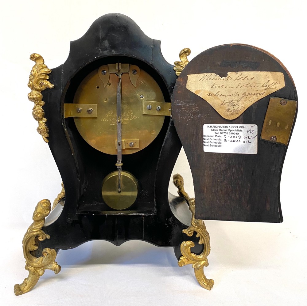 An antique ebonised wooden mantel clock featuring ornate cast ormolu mounts, a white dial with Roman numerals, and scrolled gilt brass feet. View XII