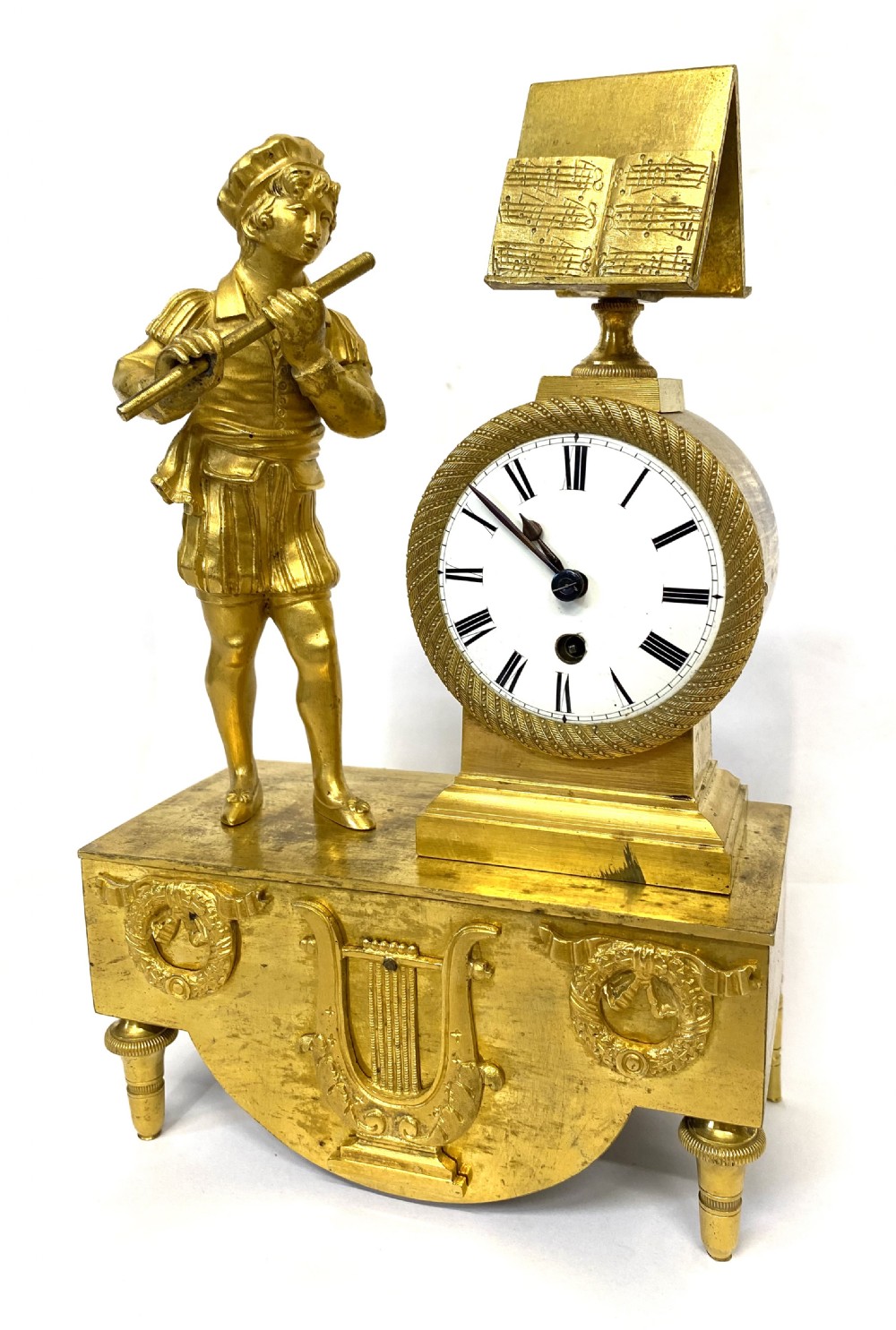 An antique gilt bronze mantel clock featuring a standing figure of a flute player beside a circular white dial topped with a music stand.