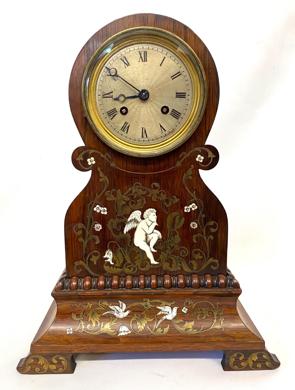 An antique French rosewood mantel clock featuring detailed brass foliage marquetry, bone cherub inlay, and a circular Roman numeral dial.