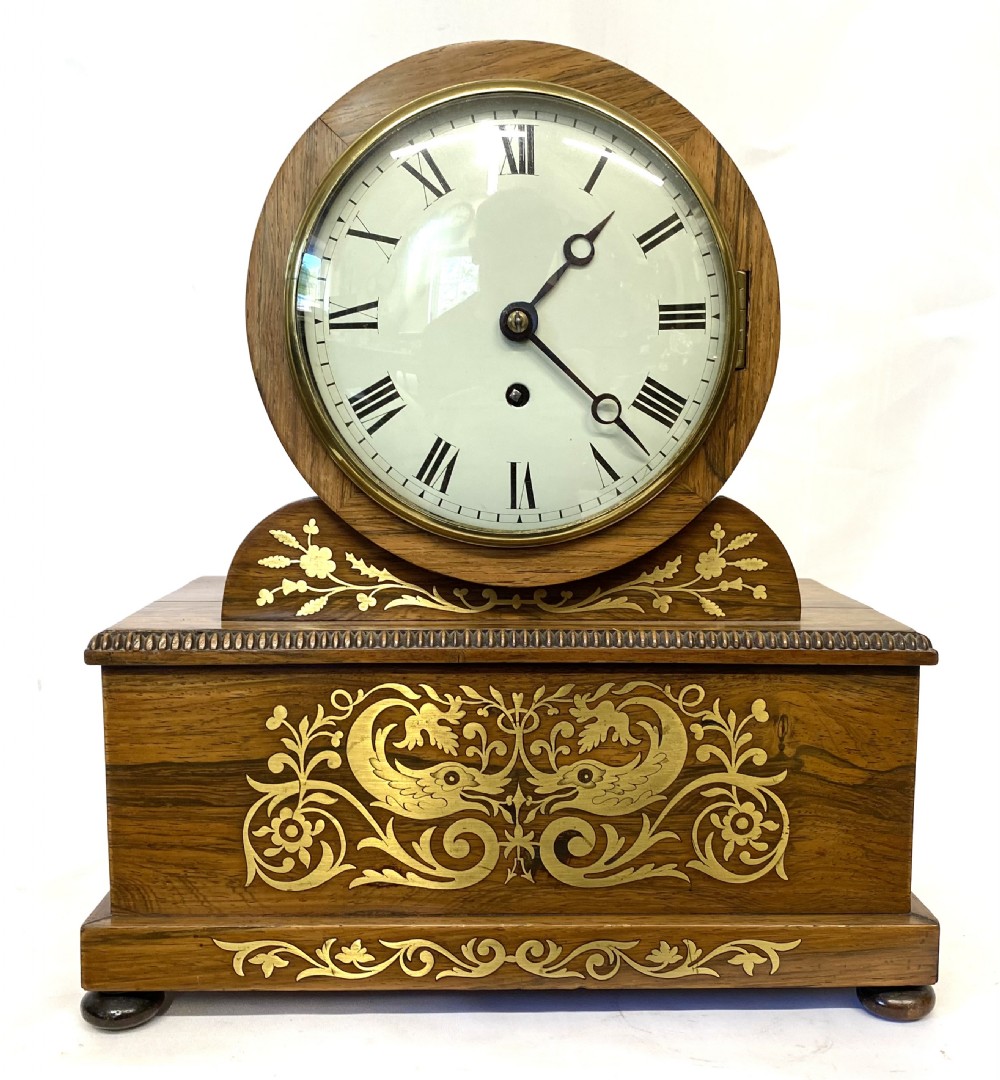 An antique Regency drumhead mantel clock constructed from polished timber with intricate brass floral inlay, a white dial with Roman numerals, and brass ball feet.