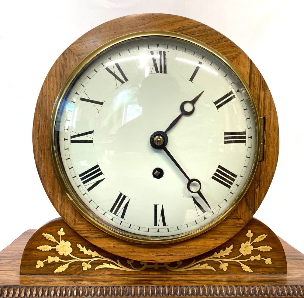 An antique Regency drumhead mantel clock constructed from polished timber with intricate brass floral inlay, a white dial with Roman numerals, and brass ball feet. View V
