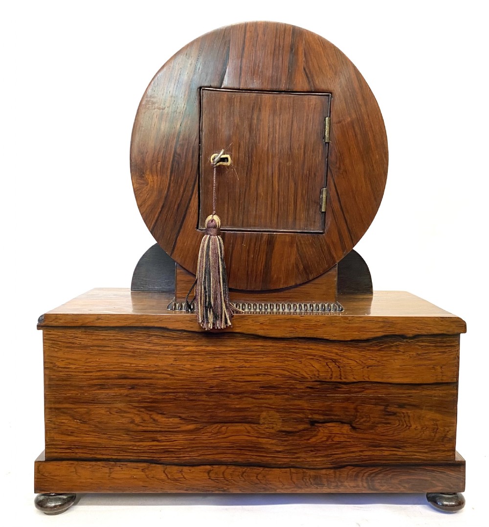 An antique Regency drumhead mantel clock constructed from polished timber with intricate brass floral inlay, a white dial with Roman numerals, and brass ball feet. View IX