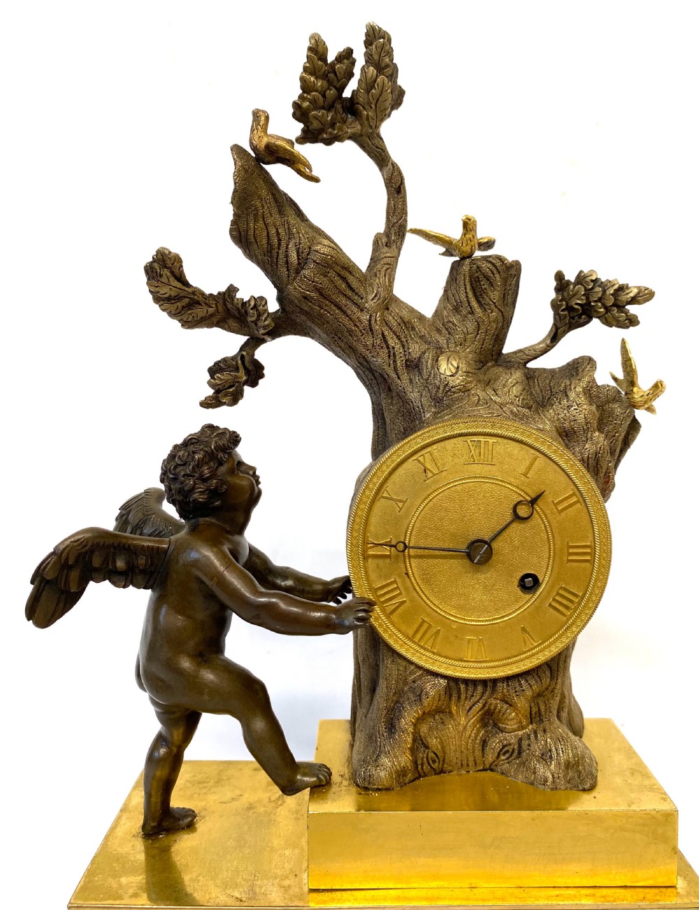 An antique gilt and patinated bronze mantel clock featuring a dark bronze cherub beside a tree stump clock housing topped with small birds. View II